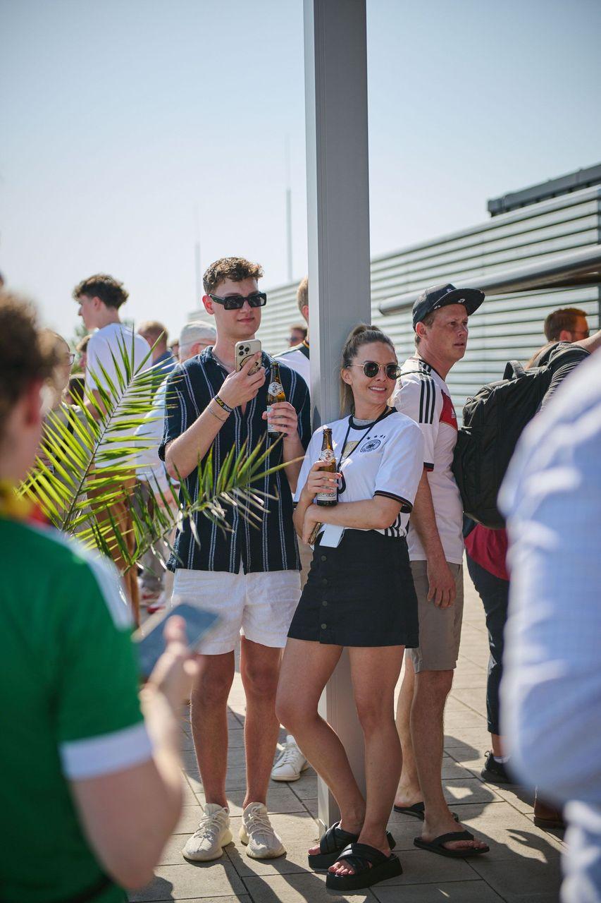 Personen auf einer Dachterrasse, die ein Deutschlandspiel der EM 2024 verfolgen; eine Frau im deutschen Trikot und ein Mann mit einem Getränk in der Hand stehen in entspannter Atmosphäre zusammen.