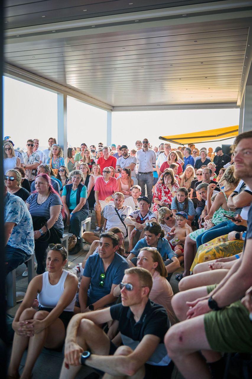 Große Gruppe von Menschen, die auf einer Dachterrasse zusammengekommen sind, um ein Deutschlandspiel der EM 2024 zu verfolgen; einige sitzen auf Bänken und dem Boden, während andere stehen.