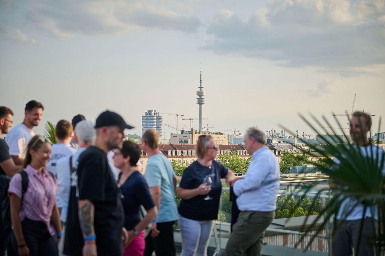 Personen auf einer Dachterrasse in München im Gespräch, im Hintergrund ist der Olympiaturm sowie die Skyline der Stadt mit Baukränen zu sehen.