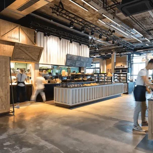 Moderner Gastronomiebereich mit industriellem Design, sichtbaren Rohren an der Decke und einer langen Verkaufstheke, hinter der Brot und Gebäck ausgestellt sind; im Vordergrund sind mehrere Personen in Bewegung.