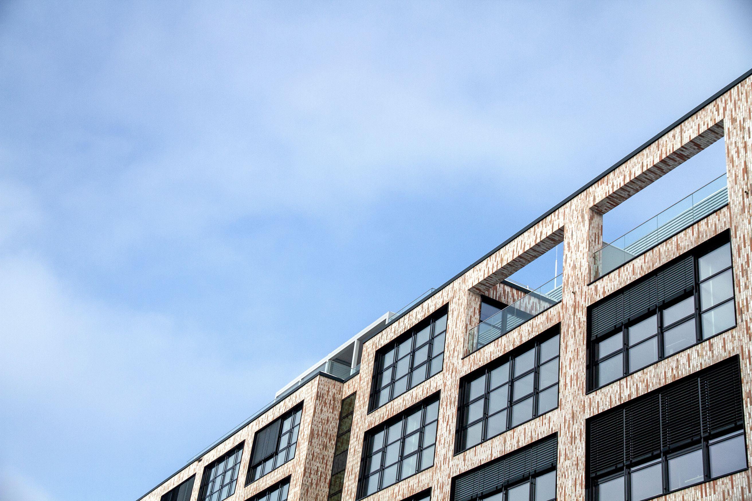 Nahaufnahme des oberen Teils des Gebäudekomplexes Neue Siederei, mit modernen Glasbalkonen und großen Fenstern, vor einem blauen Himmel.