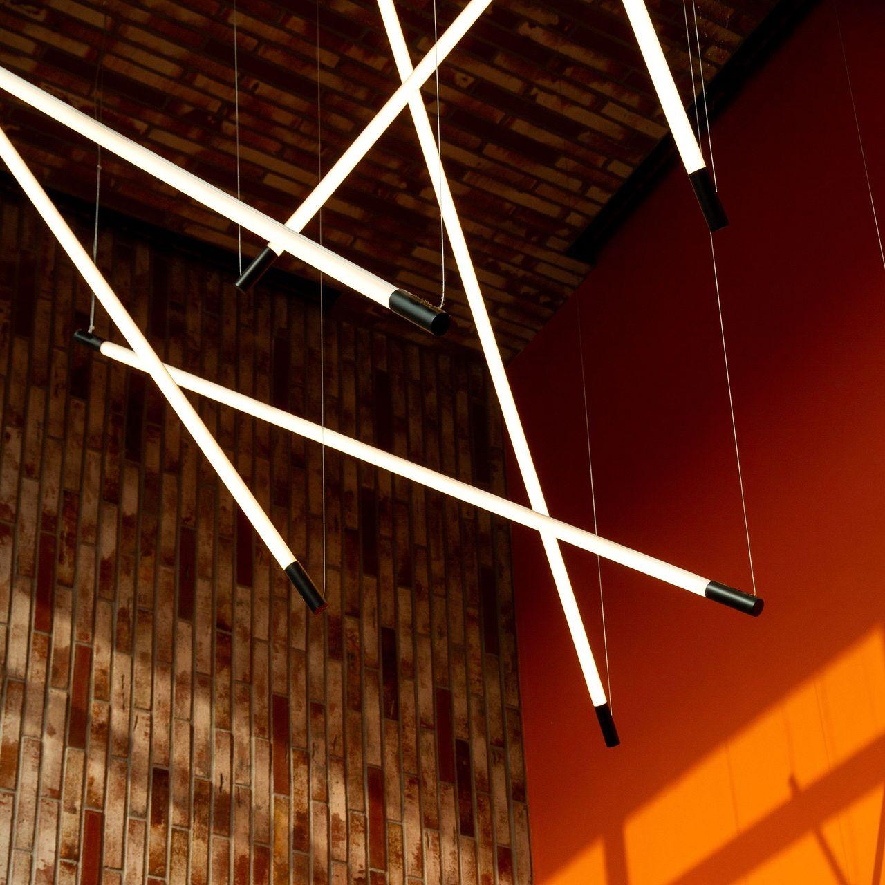 Detailaufnahme einer modernen Hängelampe in einem Innenraum mit einer rustikalen Backsteinwand und einer orangefarbenen Wand, die durch das Licht beschienen wird.