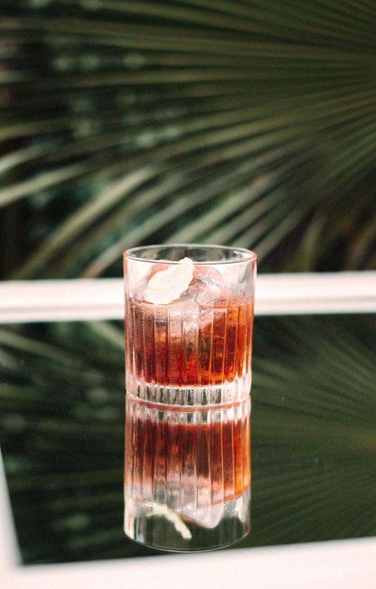 Ein roter Cocktail in einem Glas mit Eiswürfeln, auf einem glänzenden Tisch, mit Palmenblättern im Hintergrund, serviert bei einem Afterwork-Cocktailabend auf der Dachterrasse.